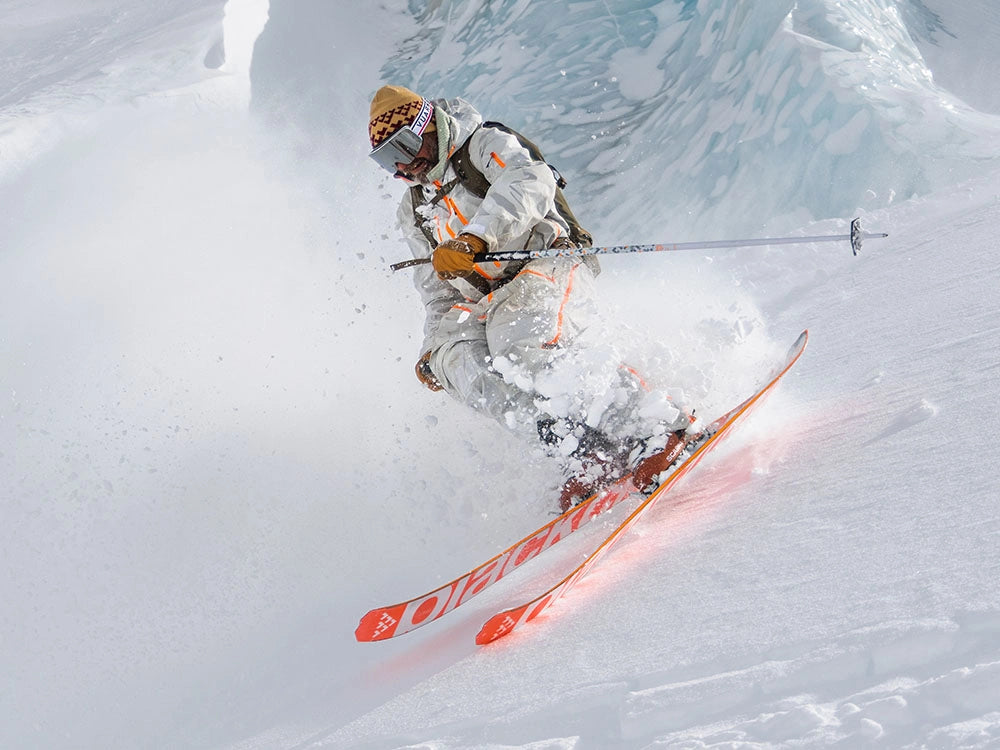 A skier in a white snowsuit with orange accents and a patterned beanie carves through deep powder snow, kicking up a spray of snow. The skier is holding ski poles and wearing orange SCARPA Maestrale ski boots.