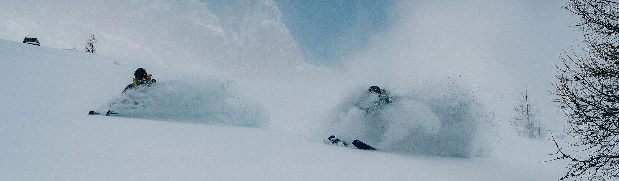 A snowy mountain slope with two skiers carving through deep powder, creating large sprays of snow, under a blue sky with sparse trees on the right.