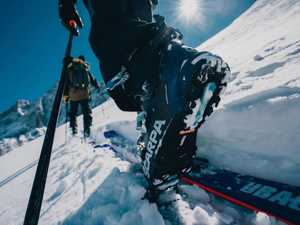 A close-up action shot of a skier skiing uphill, wearing a blue Scarpa ski boot. Another skier is visible in the background on a snowy mountain slope under a bright sun and clear blue sky.