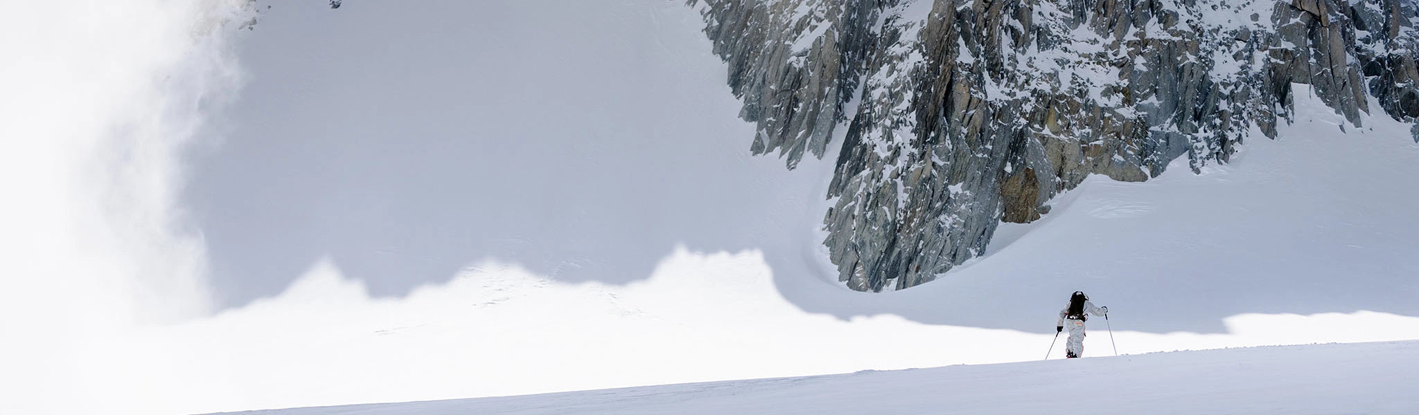 A skier stands on a snowy mountain slope beneath a steep, snow-covered rock face, with a large shadow to the left